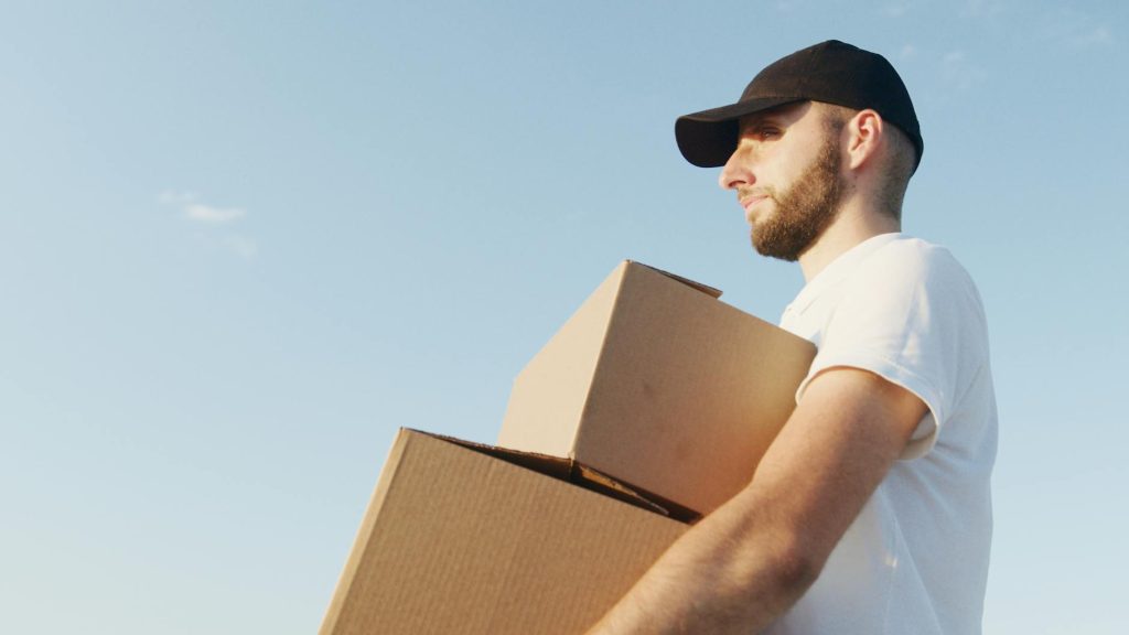 Deliveryman with beard and black hat holds boxes outdoors in daylight.