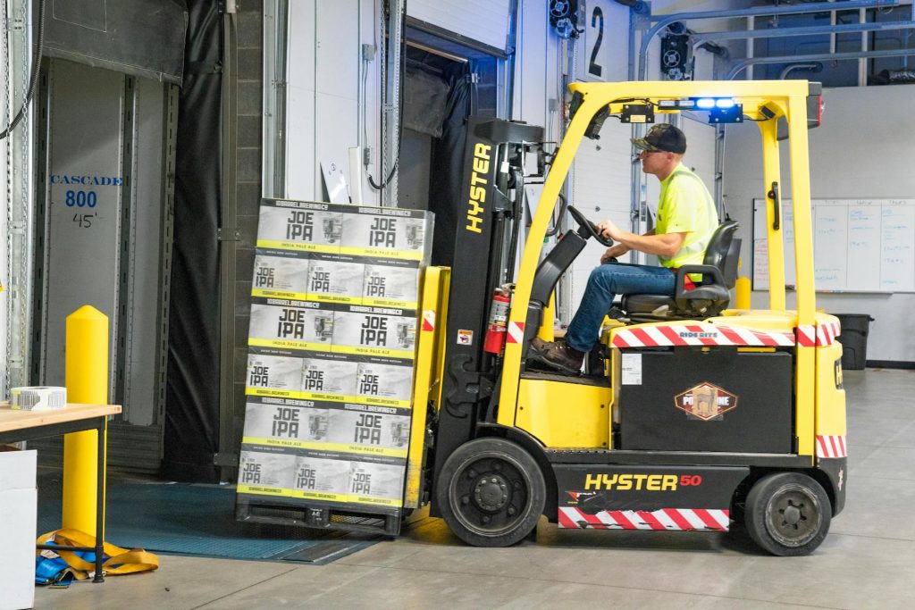 A worker drives a Hyster forklift moving Joe IPA boxes in a warehouse.