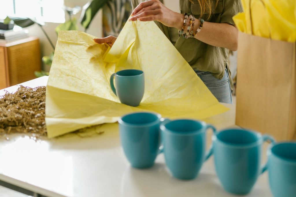 A person carefully wraps blue mugs with yellow tissue paper, showcasing packaging and preparation.
