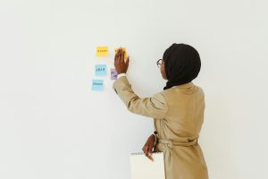 Woman in hijab organizing colorful sticky notes on a wall indoors, holding a notepad.