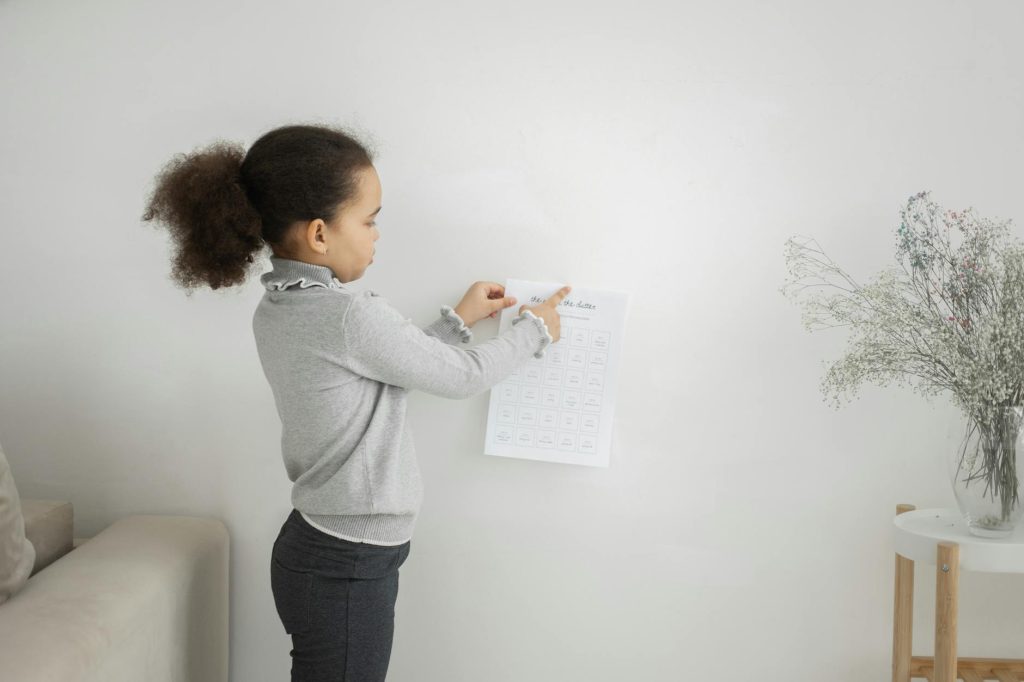 Side view of cute African American girl in casual clothes holding paper while standing near white wall in light apartment
