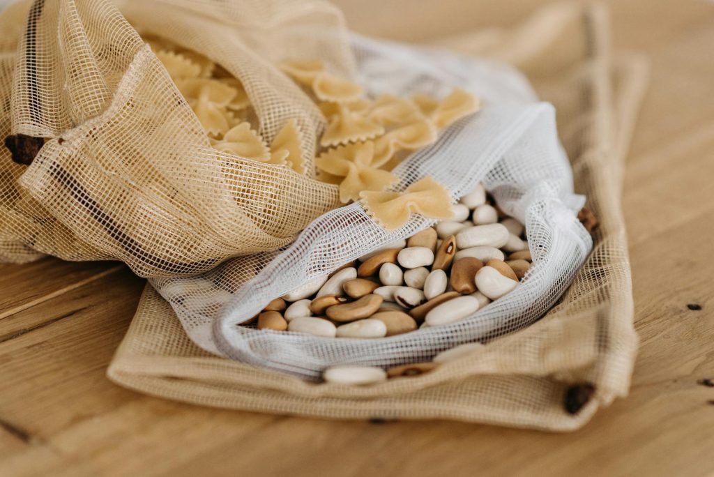 Eco-friendly bags filled with raw farfalle pasta and mixed beans on a wooden surface.