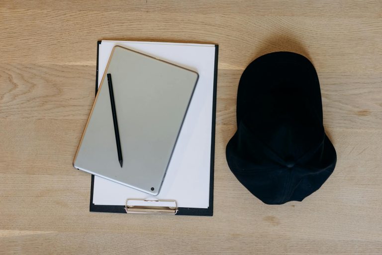 A stylish flat lay of a clipboard, tablet, and cap on a wooden desk, perfect for productivity themes.