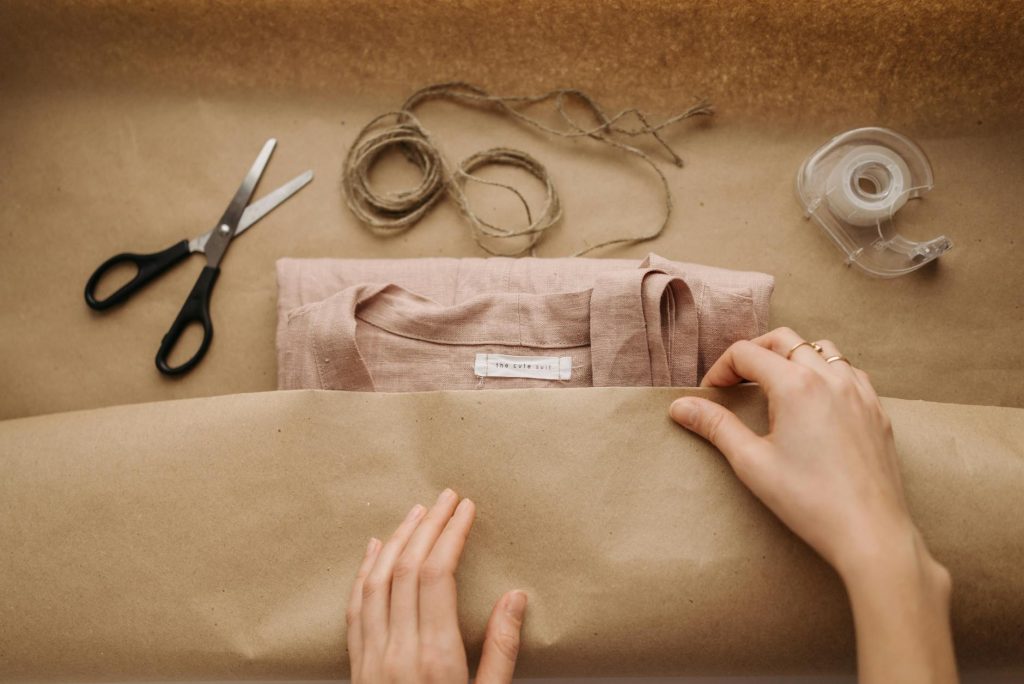 Close-up of hands wrapping a linen shirt with twine, scissors, and tape on brown paper.