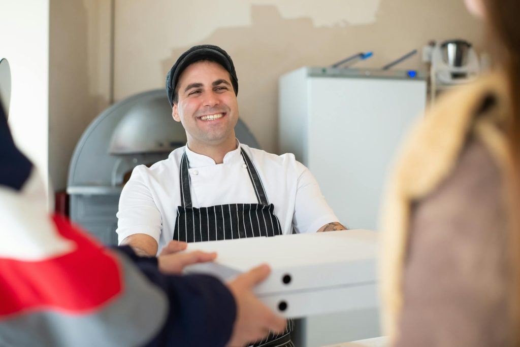 A cheerful baker in an apron hands pizza boxes to customers inside a bakery.