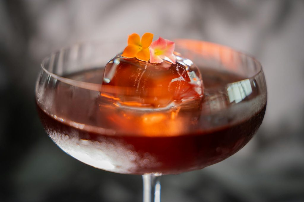 Close-up of a cocktail with floral garnish and ice sphere in a coupe glass.
