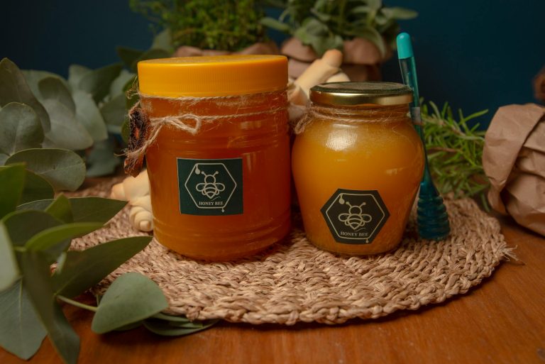 Two jars of honey on a woven mat surrounded by eucalyptus leaves and herbs.