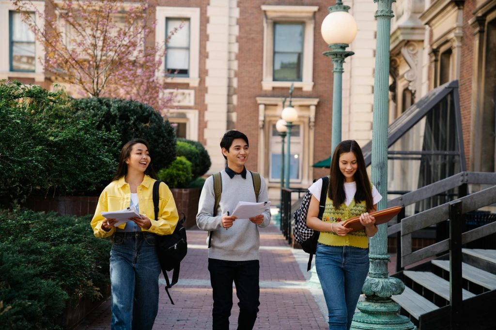 Three young adults walking and talking on a sunny campus day, holding notebooks and backpacks.