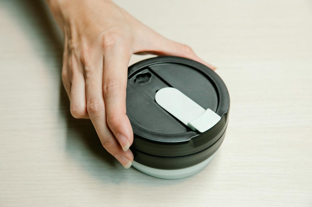 Close-up of a hand holding a black reusable coffee cup lid on a light surface.