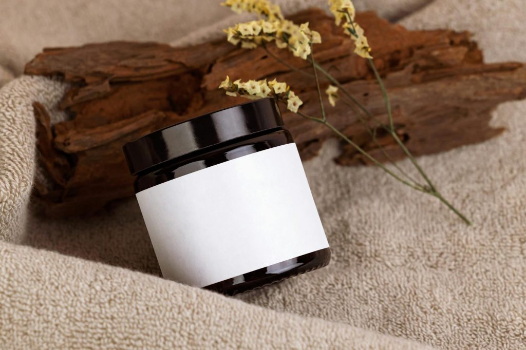 A minimalist black cosmetic jar with blank label on a natural textured towel background, accented by dried flowers and wood.