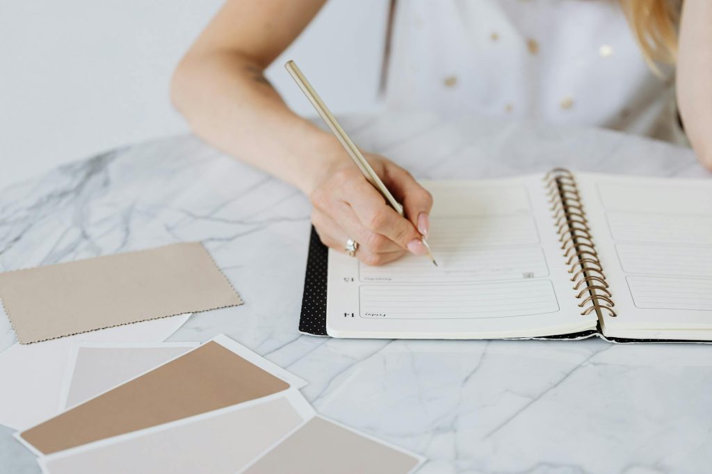 Close-up of a woman writing in a planner with color swatches on a marble table.