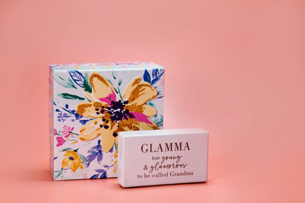 A close-up of a floral gift box and a block with the message 'Glamma' on a peach background, perfect for Mother's Day.