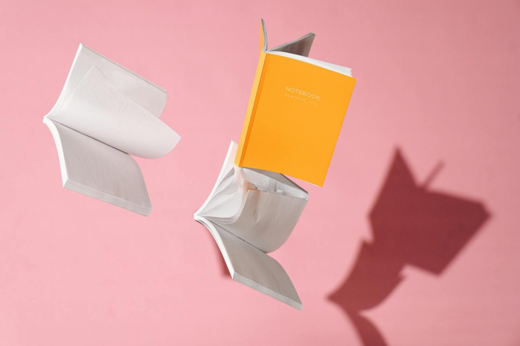 Notebooks fly against a pink backdrop, casting dynamic shadows.