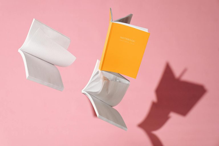 Notebooks fly against a pink backdrop, casting dynamic shadows.