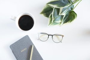 A minimalist workspace featuring a coffee cup, notebook, and eyeglasses on a white desk.