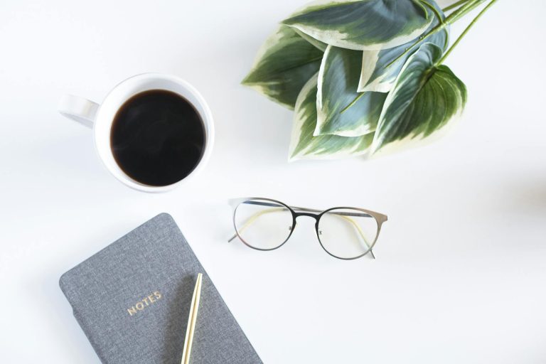 A minimalist workspace featuring a coffee cup, notebook, and eyeglasses on a white desk.