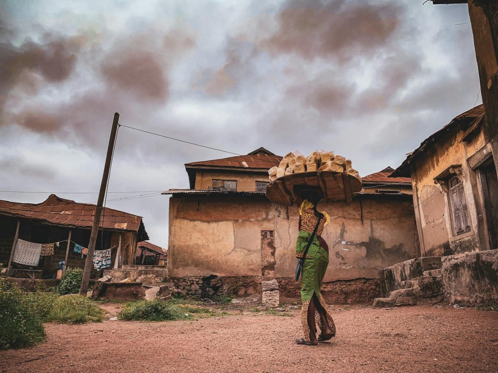 A rural village scene featuring a woman carrying goods on her head in traditional attire.