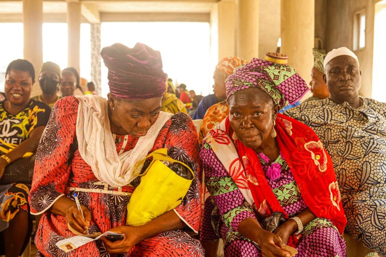 A community of adults in colorful traditional clothing gathered indoors with a mix of emotions.