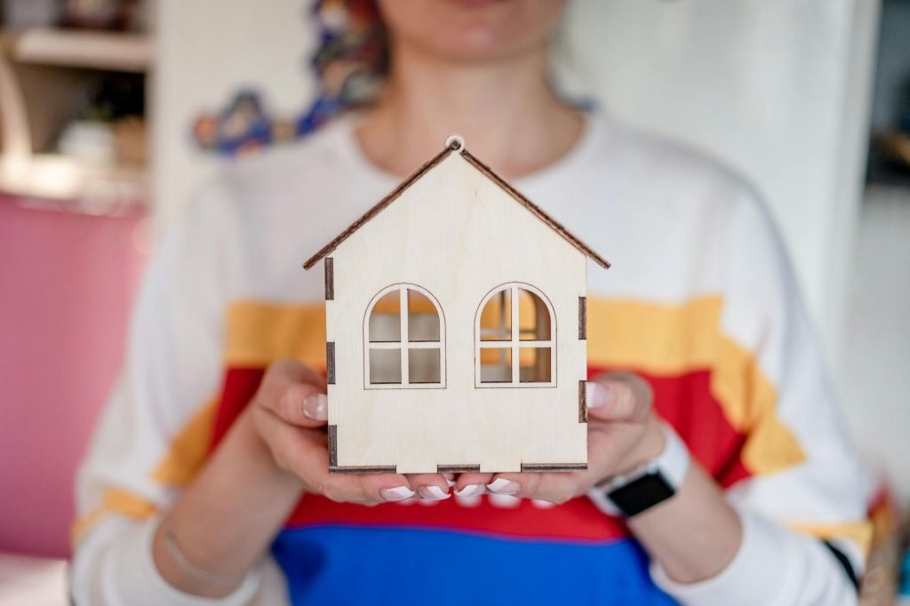 A detailed view of a woman's hands holding a handmade wooden house model indoors.