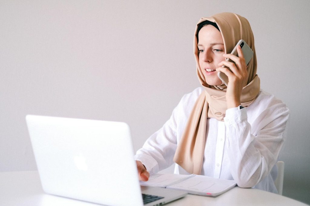 Businesswoman in hijab multitasking with laptop and phone in a home office setting.