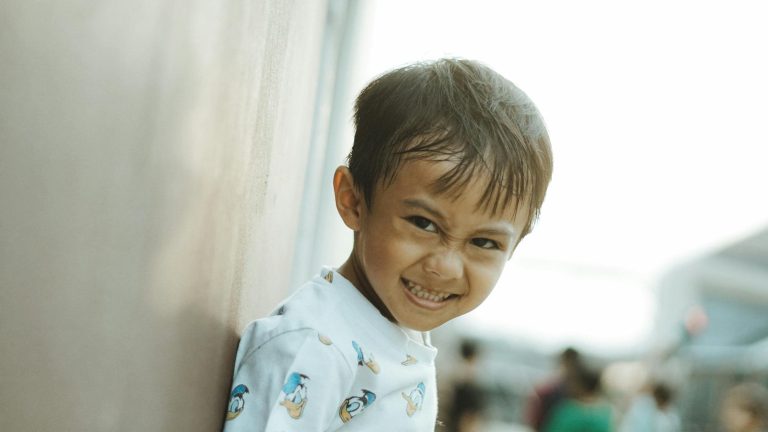 A happy child smiling brightly while leaning on a wall outdoors in West Java, Indonesia.