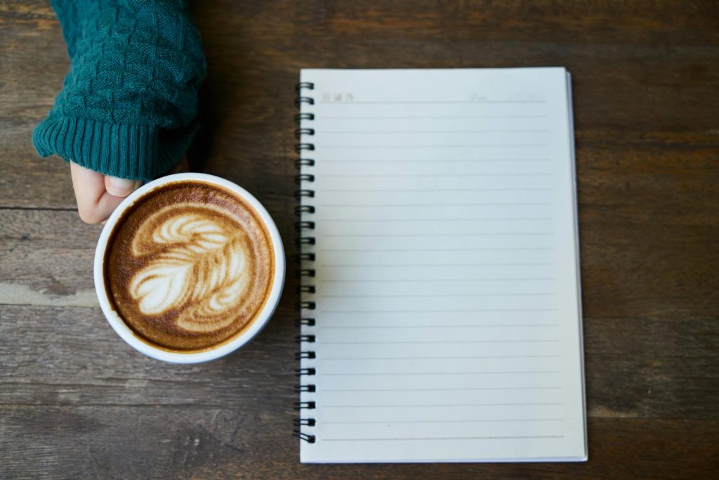 Flat lay of a coffee cup with latte art next to a blank notebook on a wooden table.