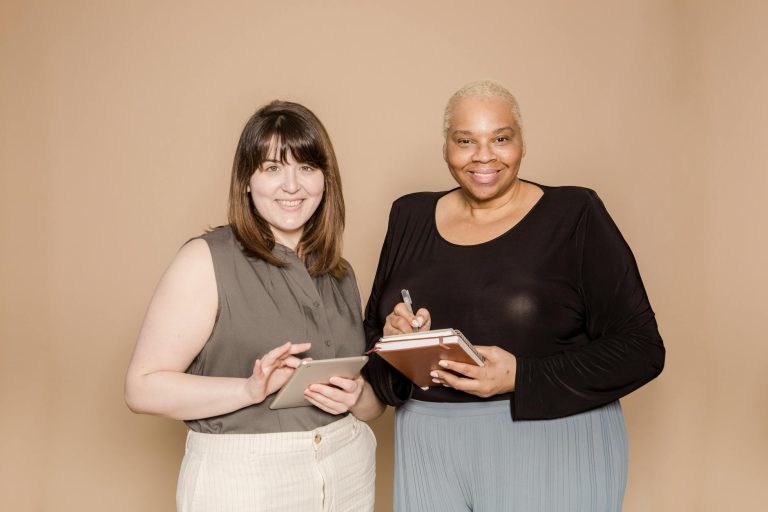 Cheerful Asian woman surfing internet on tablet near happy overweight African American female colleague with notebooks while looking at camera on beige background in studio
