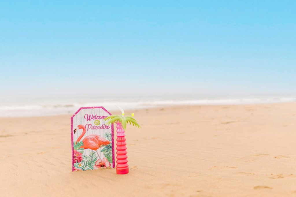 Bright beach scene with a "Welcome to Paradise" sign and a tropical drink on the sand.