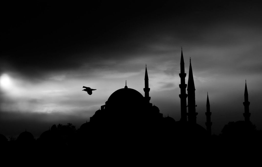 A black and white photo of a mosque silhouette with a bird flying during twilight.