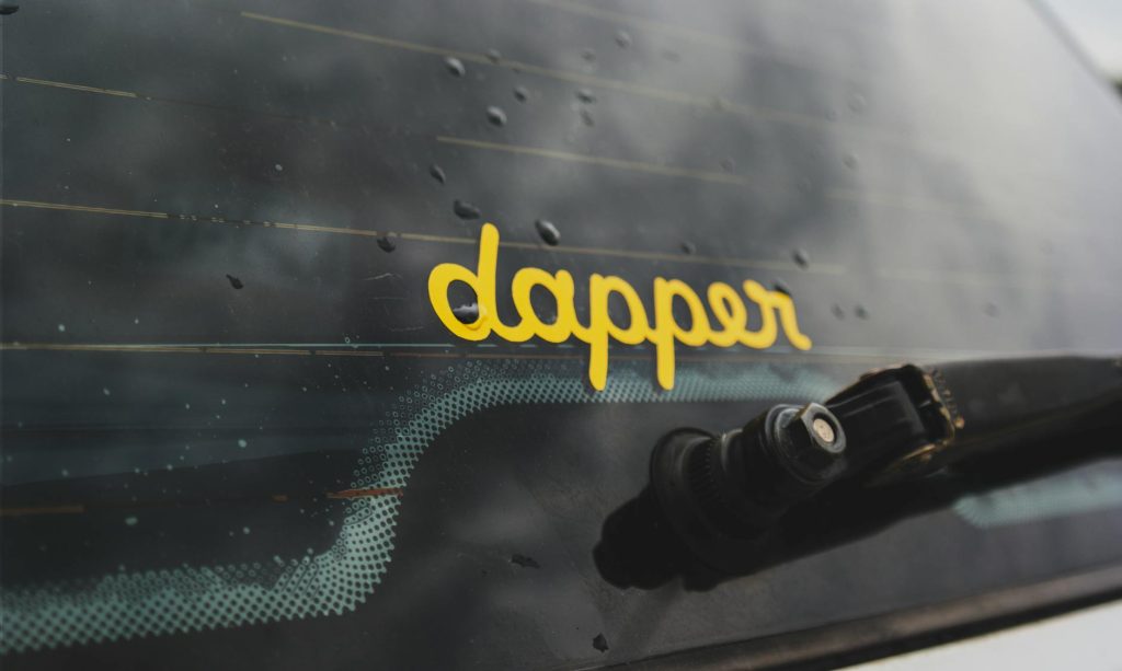 A close-up of a vehicle rear window featuring a yellow 'dapper' sticker and water droplets.