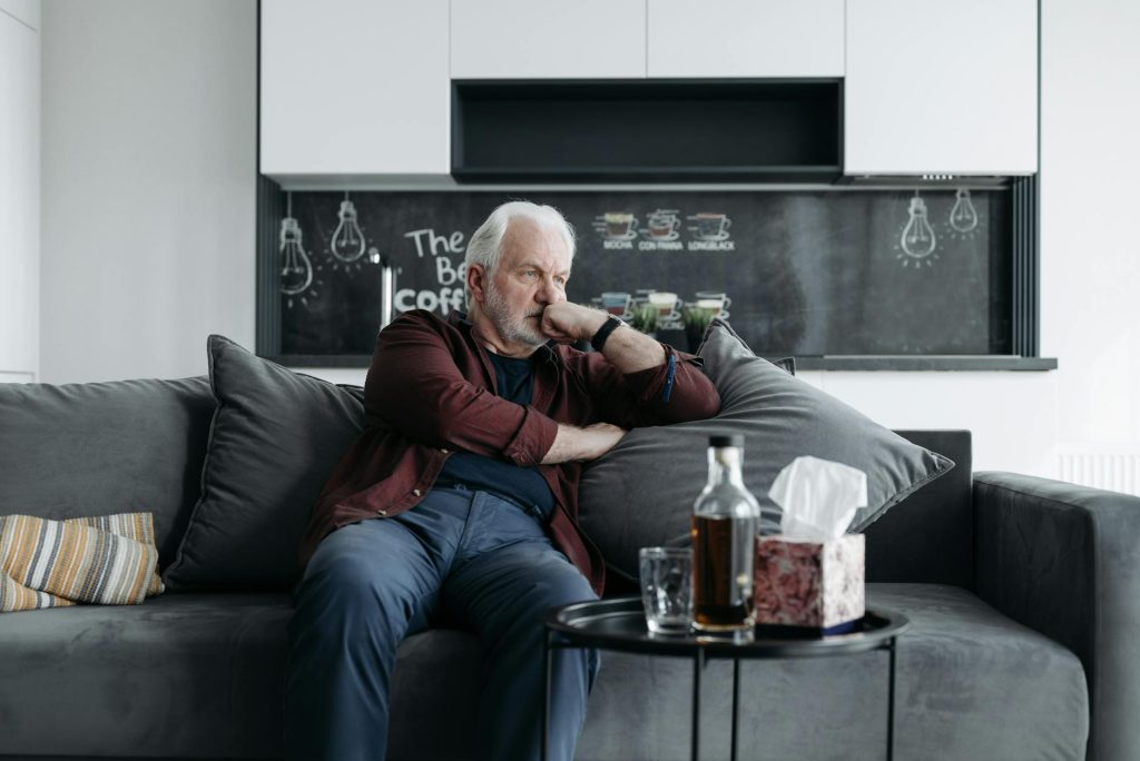 Contemplative elderly man sitting on a couch with drinks and tissues nearby, indoors.