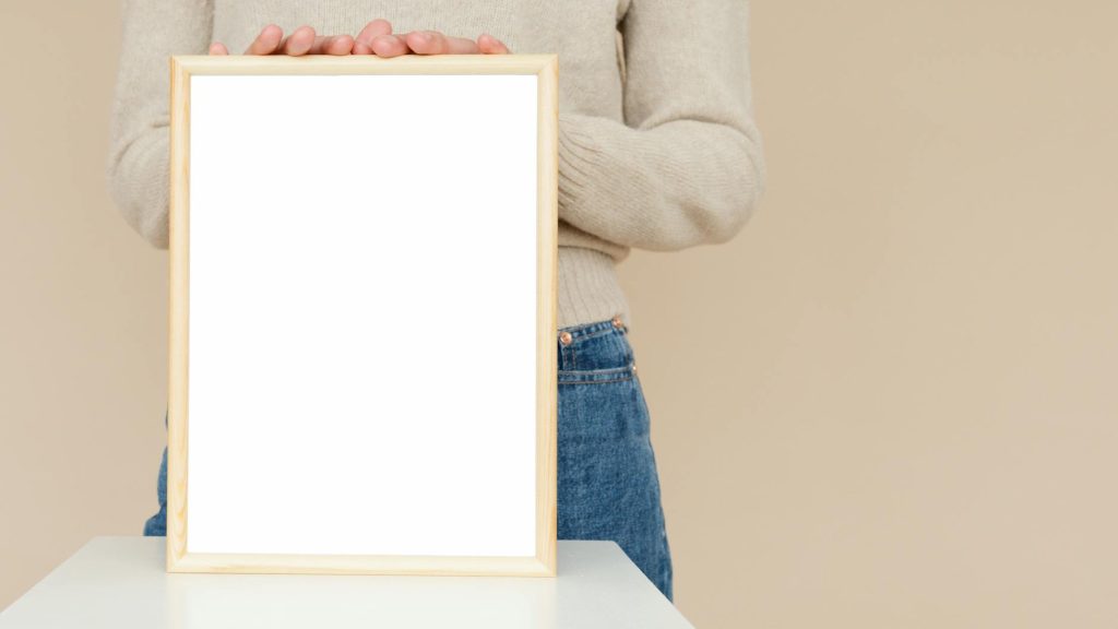 Person holding a blank white wooden frame indoors against a neutral background.