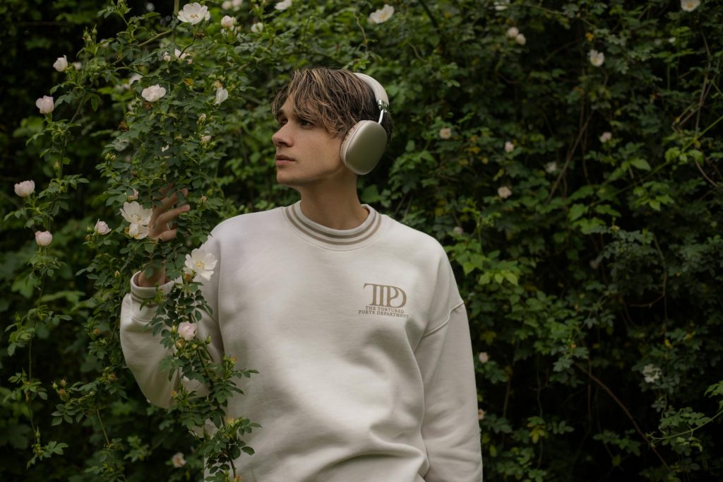 A young man wearing headphones and a white sweatshirt stands amidst lush greenery and flowers.