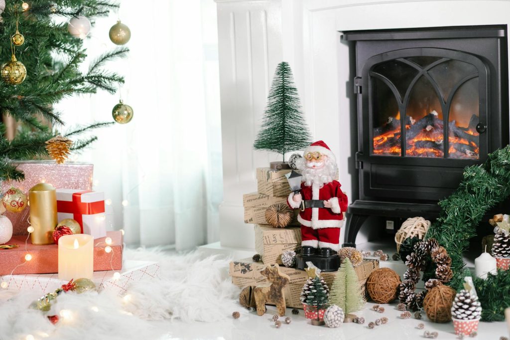 Cozy light room with gift boxes under Christmas tree and Santa Claus near holiday decorations placed on white cozy fabric at home with fireplace and burning candles
