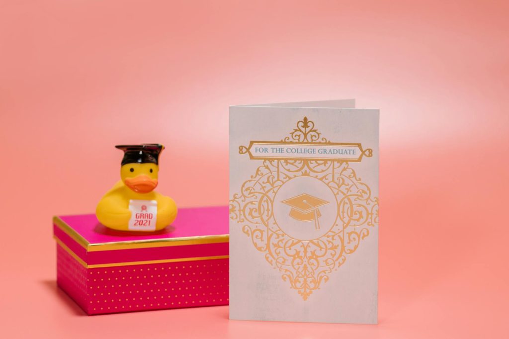 Studio shot of a graduation card, rubber duck, and gift box on a pink background.