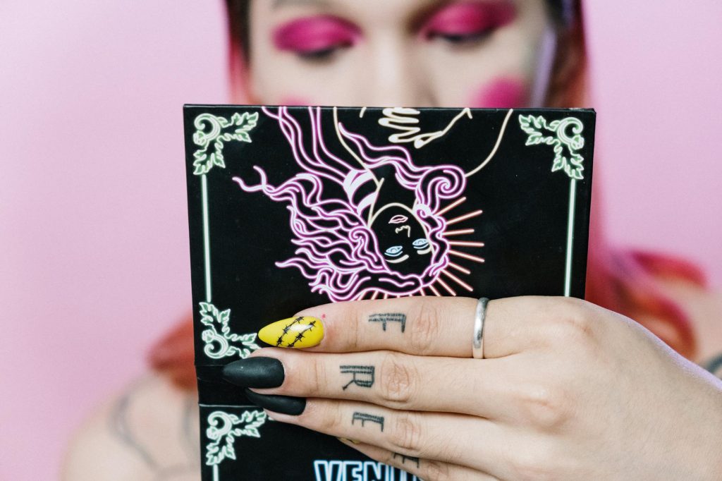 Close-up of a tattooed hand holding an artistic makeup palette with vibrant pink eye makeup in the background.