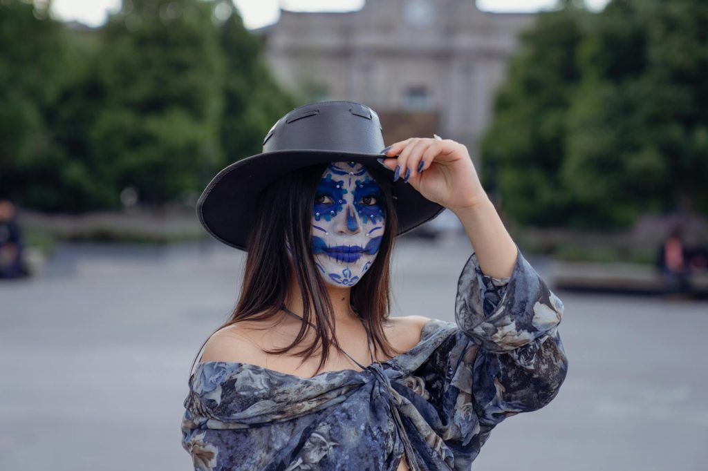 A woman wearing sugar skull makeup and a hat poses outdoors, capturing cultural artistry.
