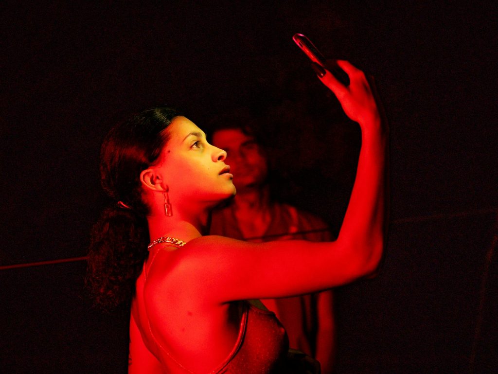 Young woman illuminated by red light, looking at phone in a moody, cinematic setting.