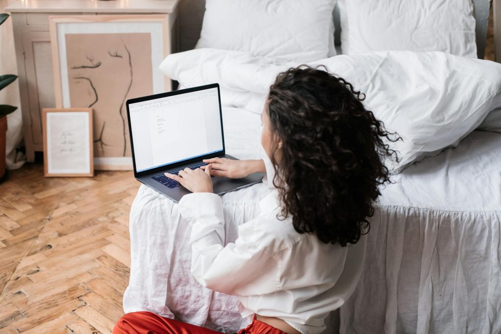 A woman works on her laptop while sitting on the bedroom floor, creating a comfortable home office environment.