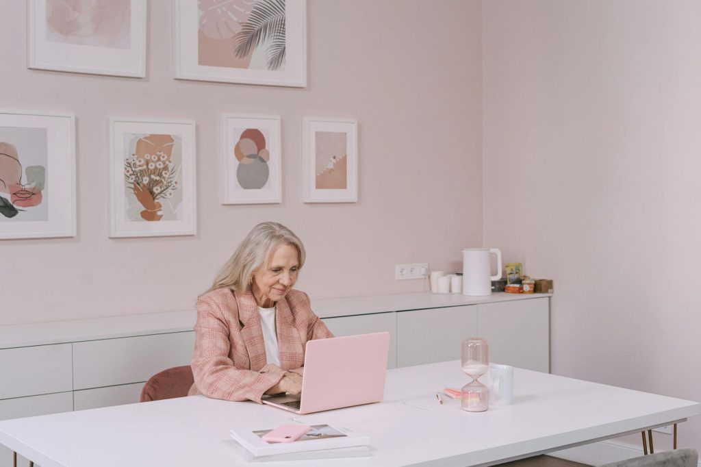 Elderly woman in pink blazer uses laptop in modern home office setting, smiling and engaged.