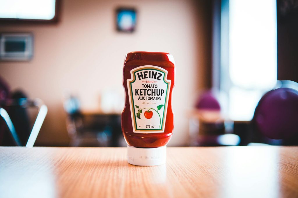 A Heinz Tomato Ketchup bottle on a wooden table with blurred background indoors.