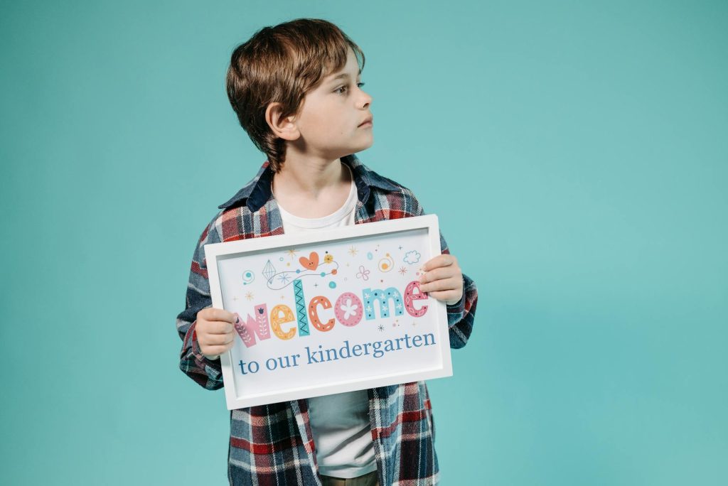 Adorable child holding a 'Welcome to our kindergarten' sign against a turquoise background.