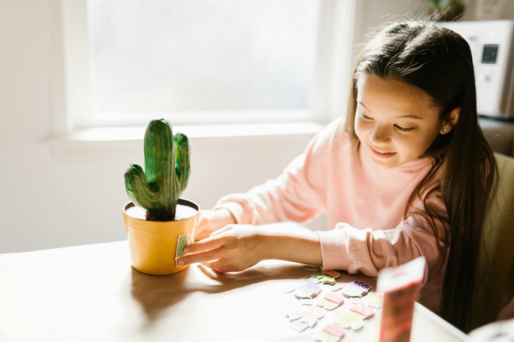 Young girl decorating a cactus pot with stickers in a bright, sunlit room, showcasing creativity and joy.