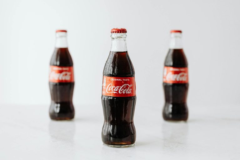 Modern glass bottles of cold soda with red label placed on white reflective surface