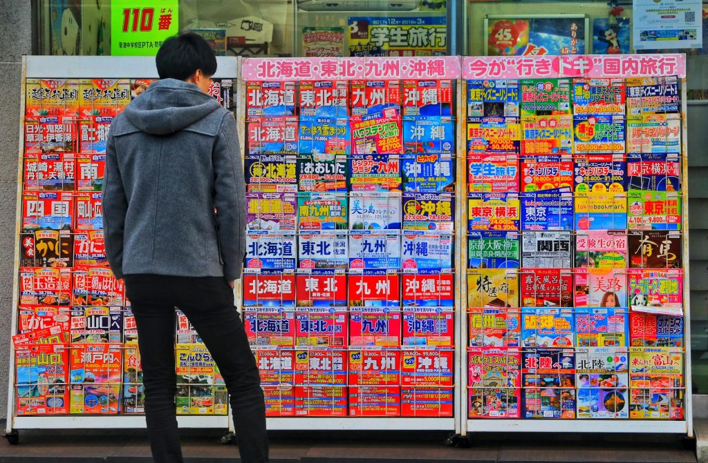 A man browsing a vibrant magazine stand on a street in Otsu, Japan, showcasing colorful travel options.