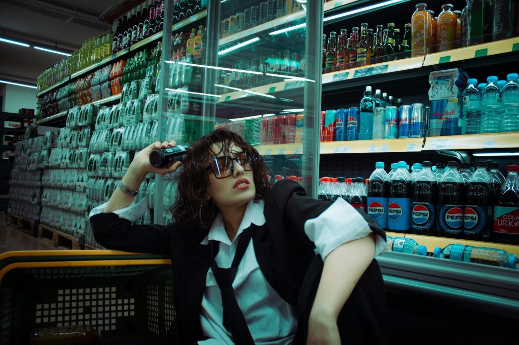Trendy woman in supermarket with sunglasses and blazer, sitting near drink shelves.