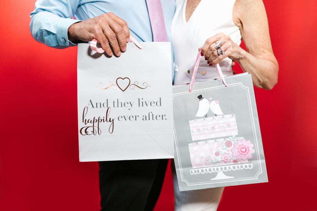 Senior couple holding gift bags with a romantic theme, against a red background.