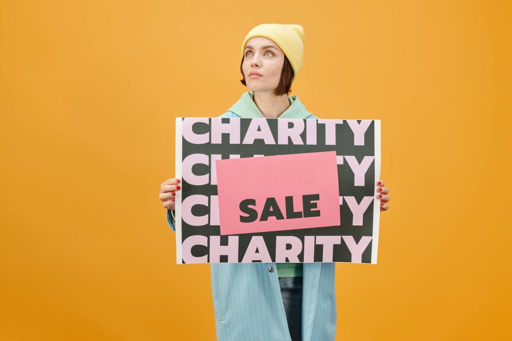 Woman in trendy outfit holding a charity sale sign against a vibrant yellow background.