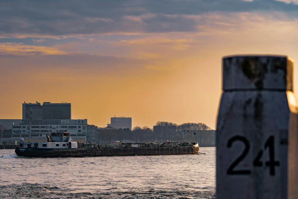 A barge travels on a river during sunset, with a cityscape in the background, creating a serene industrial scene.