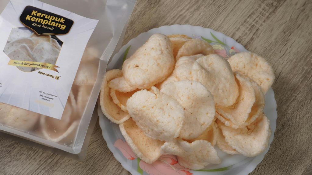 Plate of Kerupuk Kemplang crackers alongside a packaged bag on a wooden table.